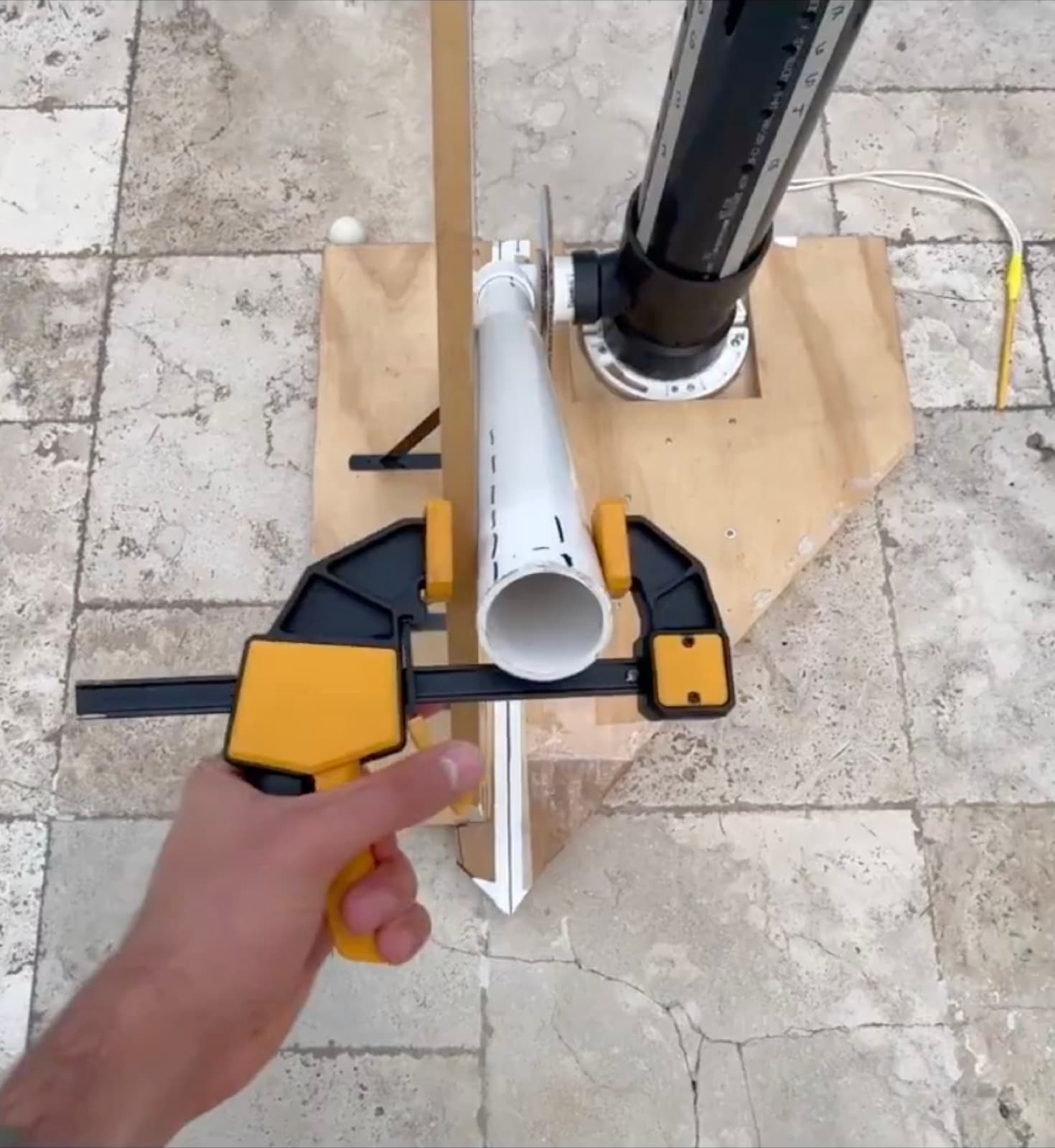 A bar clamp squeezing the white PVC barrel against a wooden upright fixed to the plywood base.