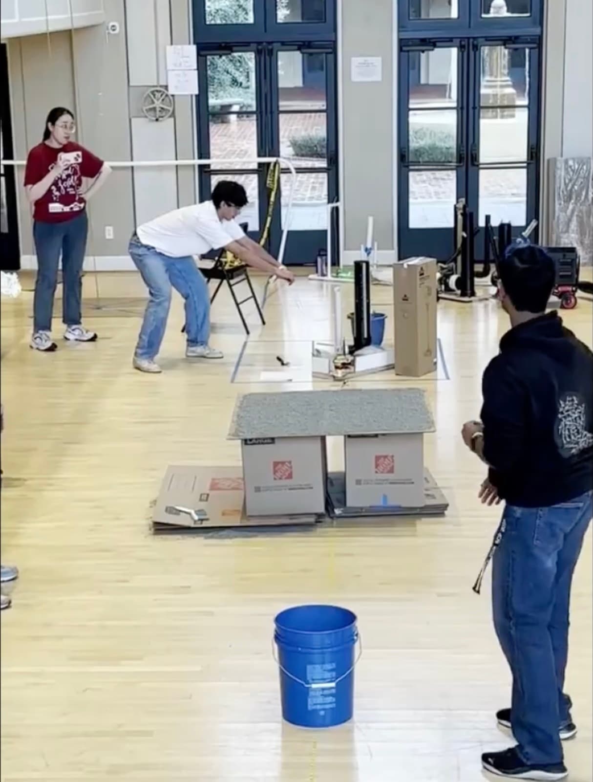 The launcher set up on a gym floor during a tournament, aimed at a raised near target resting on boxes, with a five-gallon bucket in the foreground and event supervisors watching.