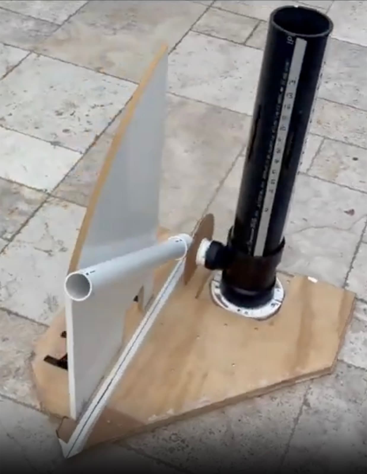 The launcher on a plywood base: a black PVC drop tube marked with a numbered height scale, a white PVC barrel angled down from its base, and a quarter-circle angle plate behind the barrel.