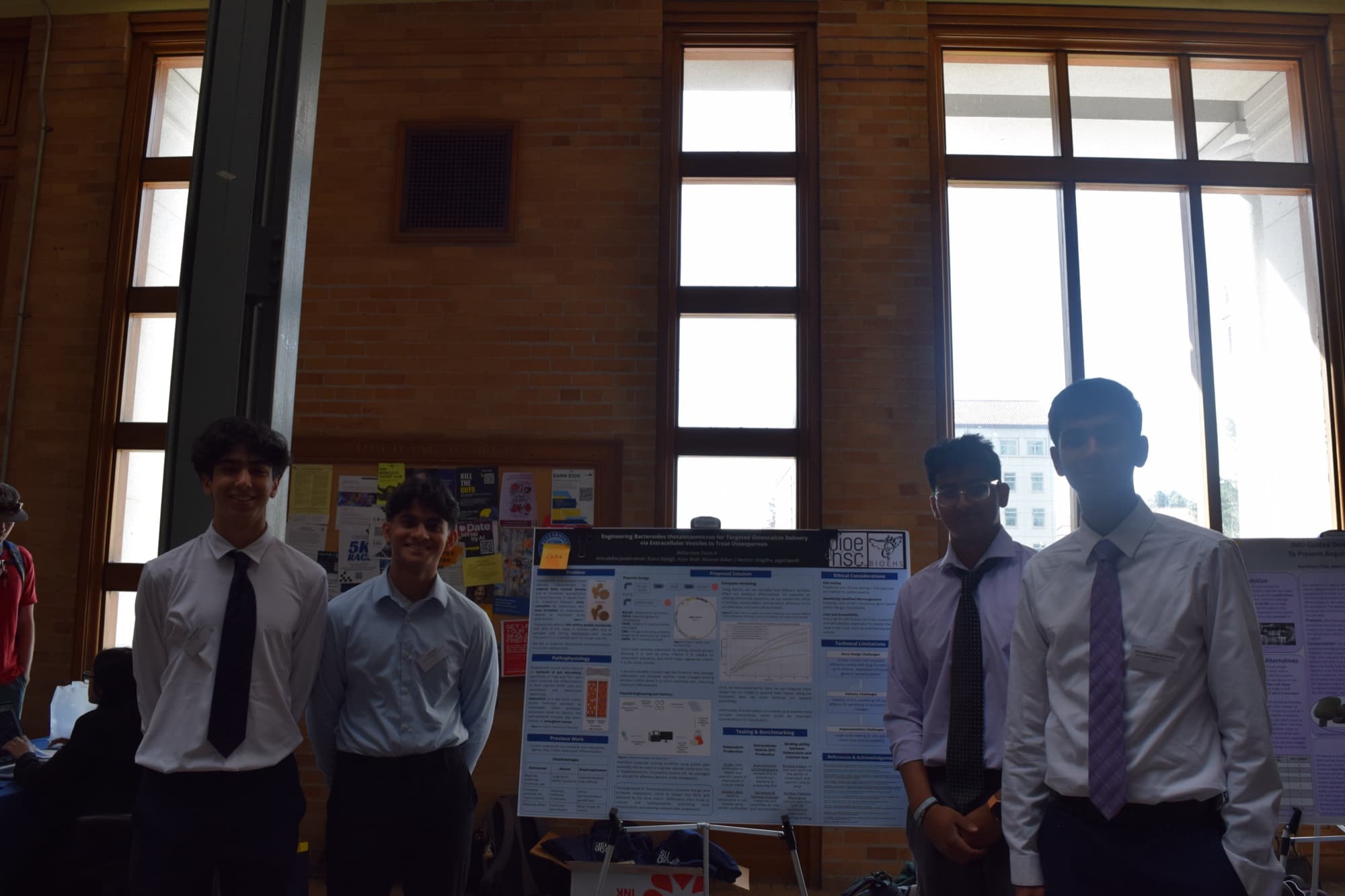 The four of us standing either side of the poster at the competition, in a hall with tall windows.